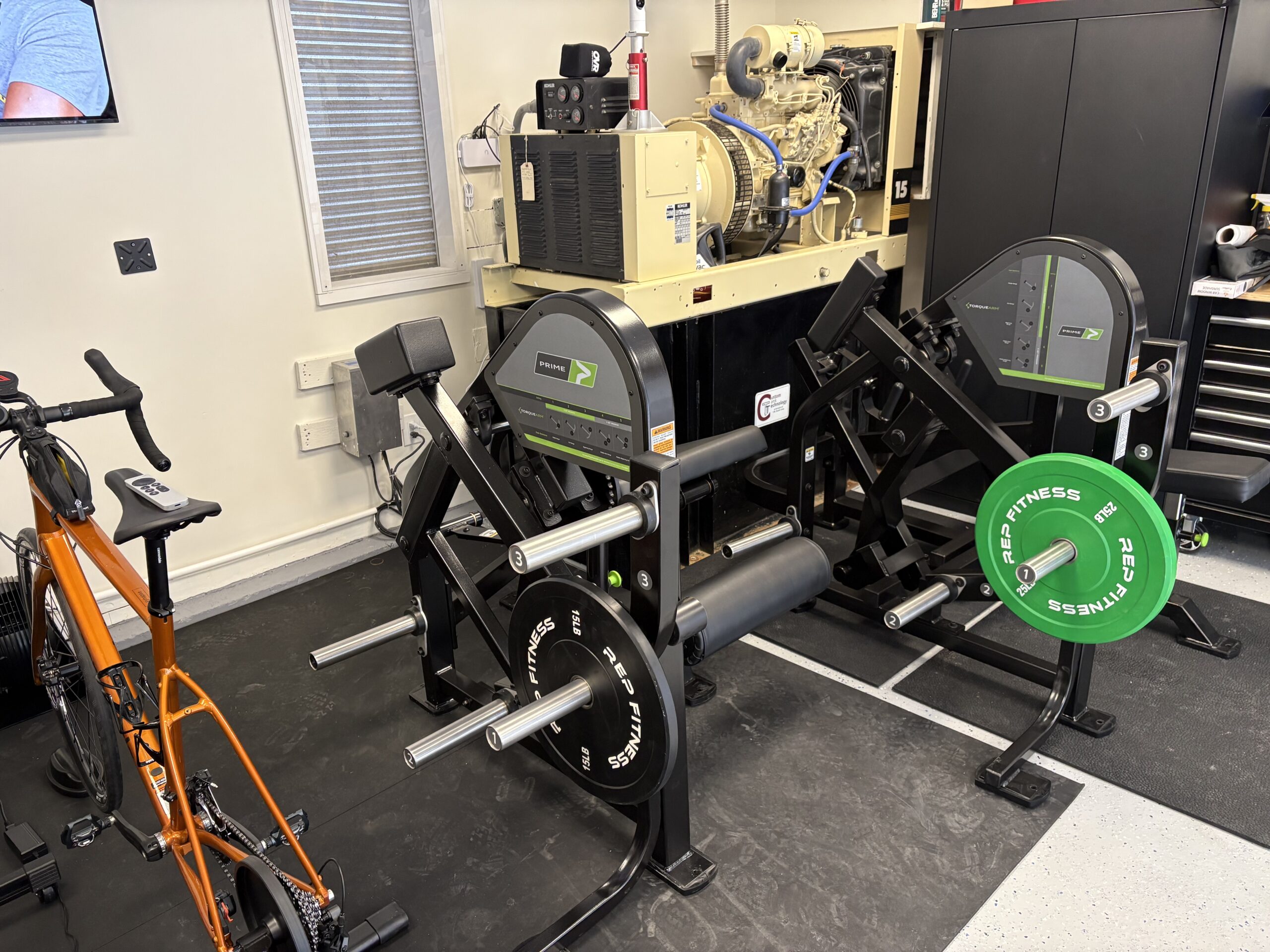 The two PRIME plate-loaded machines installed in the garage, loaded with bumper plates, with the road bike and the backup generator behind them.