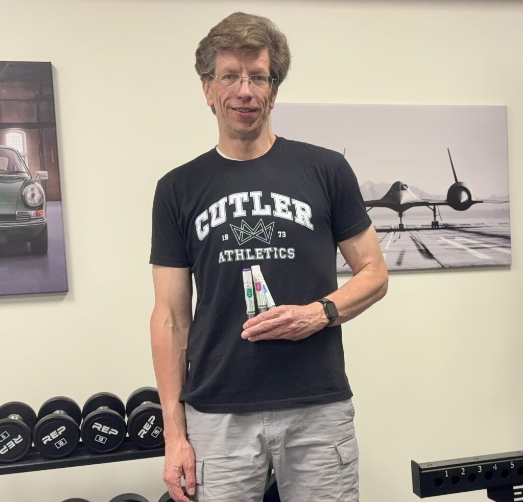 The author standing in his home gym holding two Zepbound (tirzepatide) injector pens. He is wearing a Cutler Athletics t-shirt and a smartwatch, with a rack of REP hex dumbbells and framed prints of a Porsche 911 and an SR-71 Blackbird visible behind him.