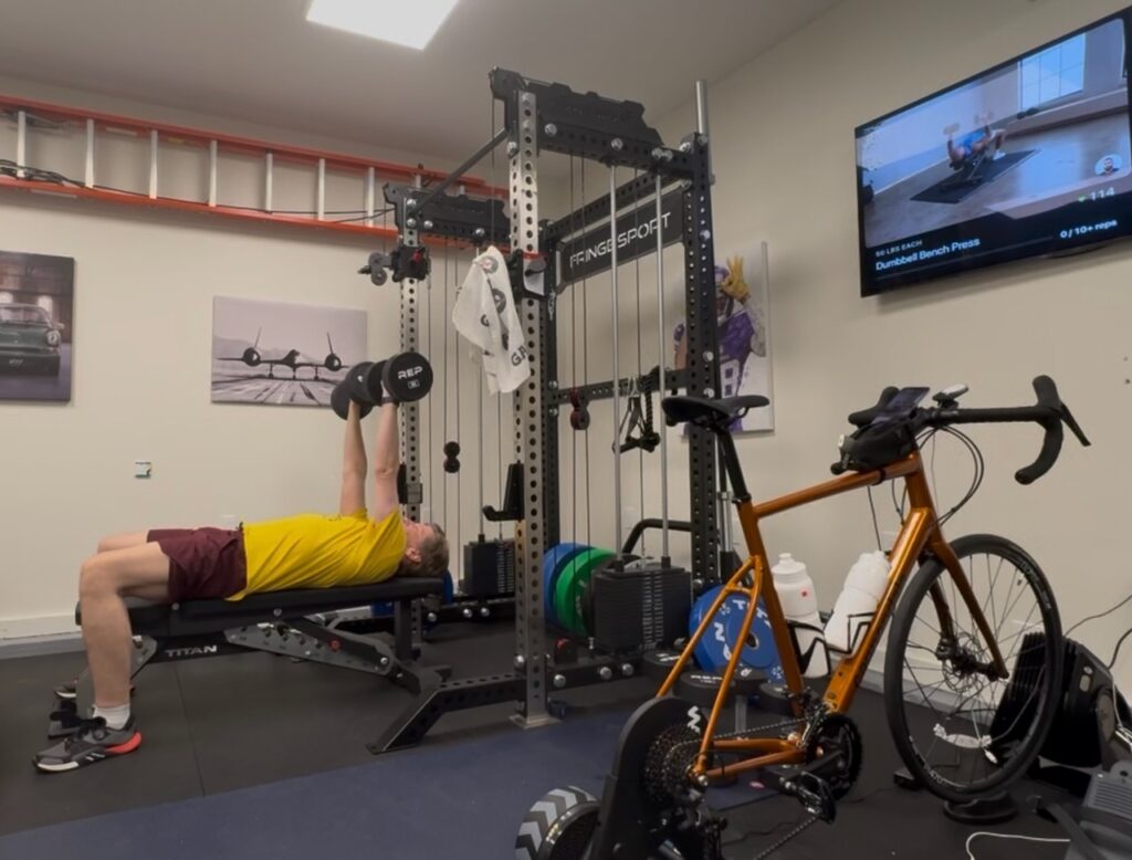 Home garage gym scene: a man does a dumbbell bench press on a Titan bench inside a Fringe Sport rack, an orange road bike sits on a Wahoo KICKR Core 2 smart trainer in the foreground, and a TV on the wall displays a workout from the Future app.