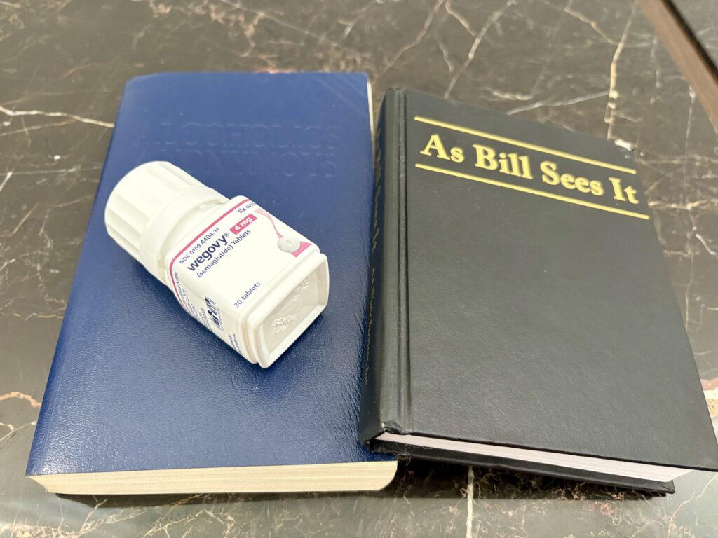 A Wegovy semaglutide pill bottle resting on a blue Alcoholics Anonymous book beside the AA daily-reflections book As Bill Sees It, on a black marble surface