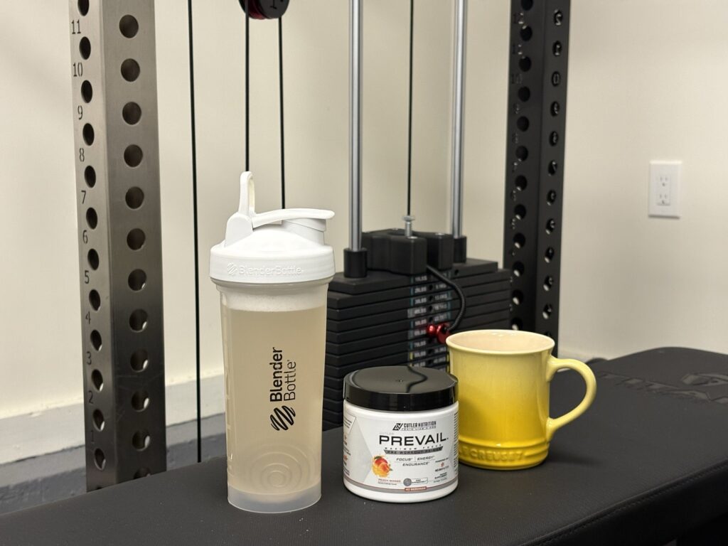 BlenderBottle shaker with mixed pre-workout, a tub of Prevail, and a yellow coffee mug on a bench in front of a garage gym rack