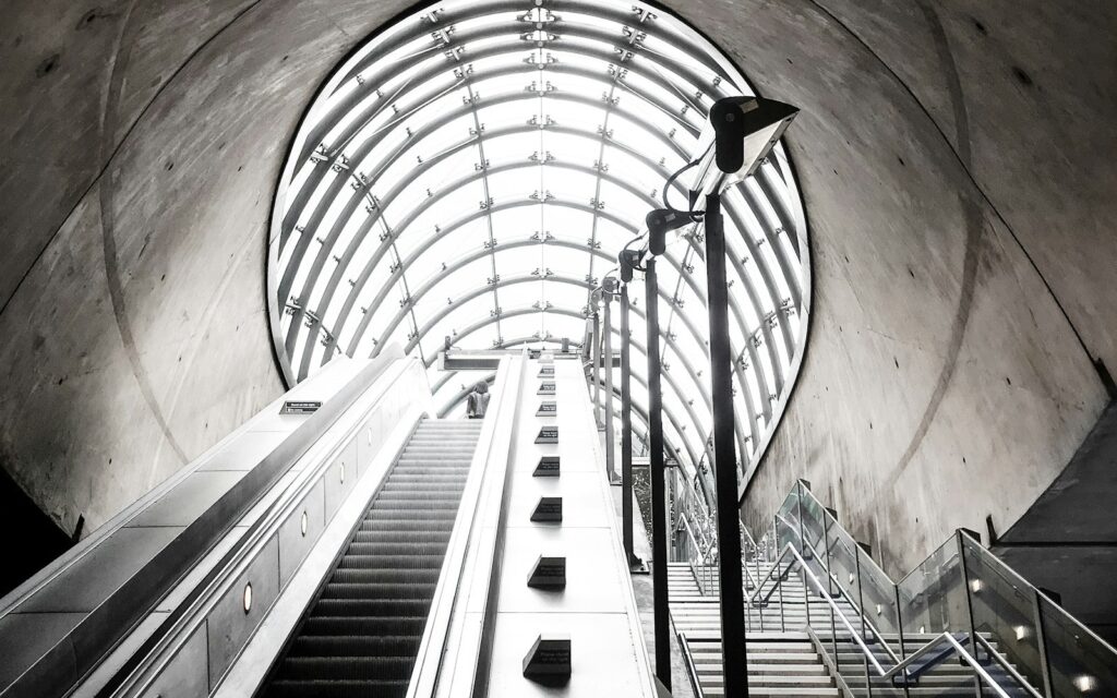 An escalator and a staircase rising side by side toward a lit glass canopy in a subway station