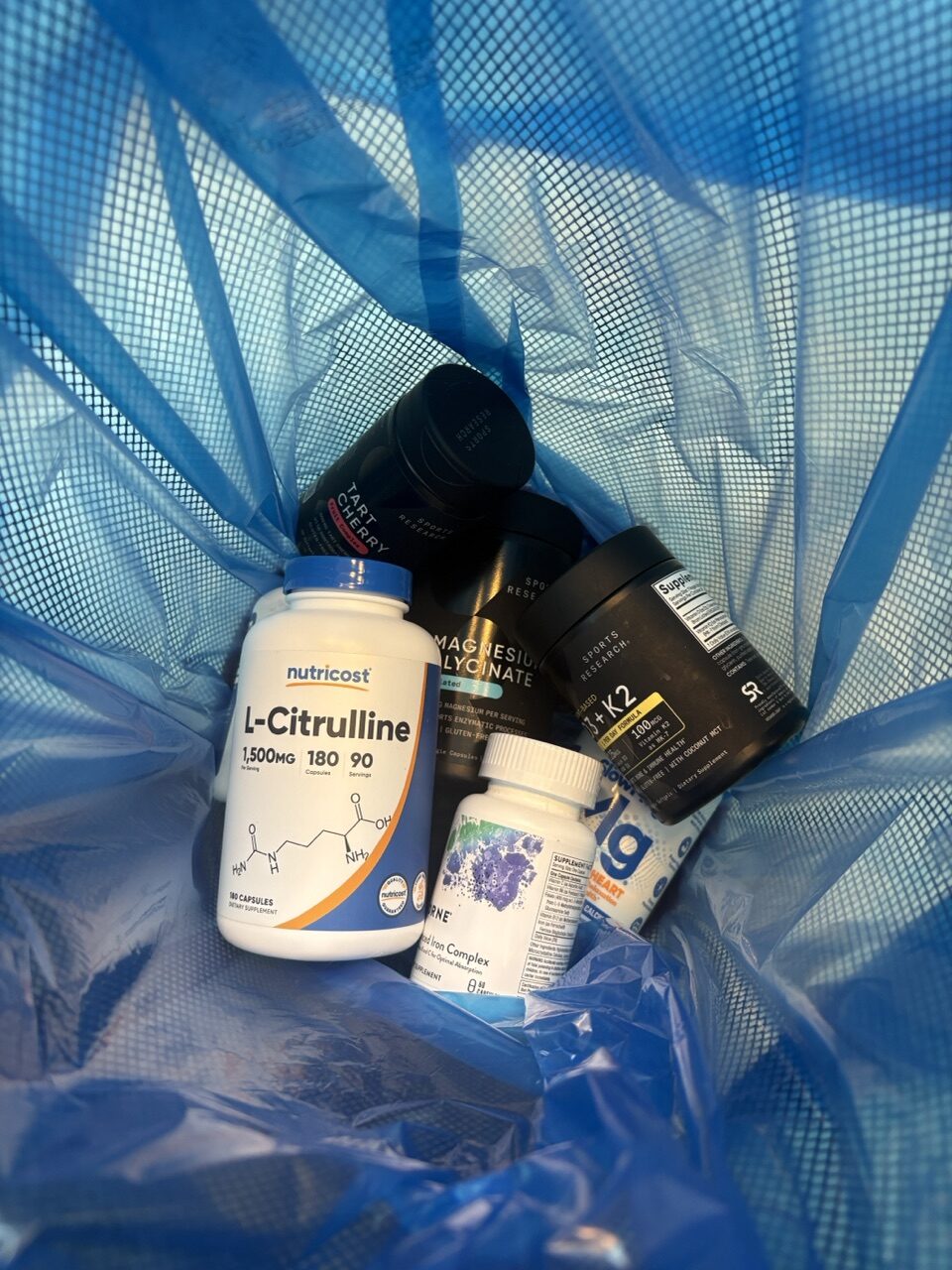 Discarded supplement bottles in a blue trash bag, including tart cherry and L-citrulline, cleared out of the cabinet