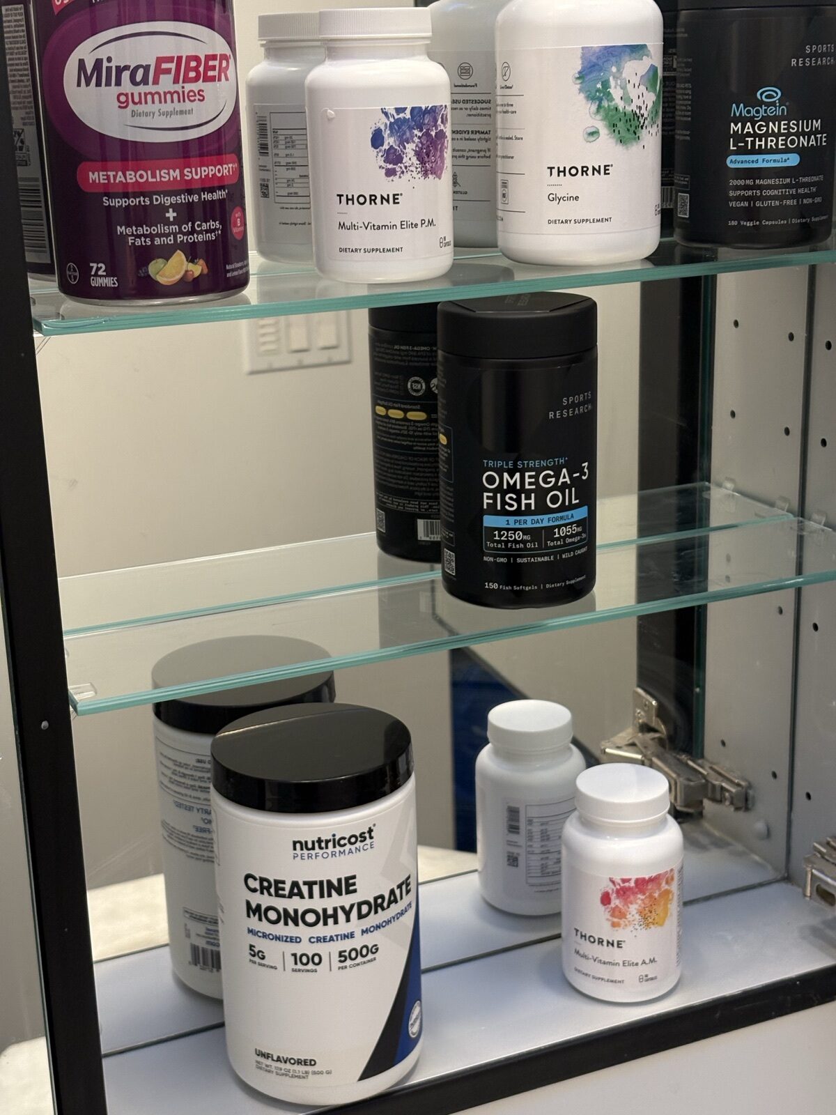 Bathroom cabinet shelf of supplement keepers: magnesium L-threonate, Thorne glycine and multivitamins, MiraFiber gummies, Omega-3 fish oil, and creatine monohydrate