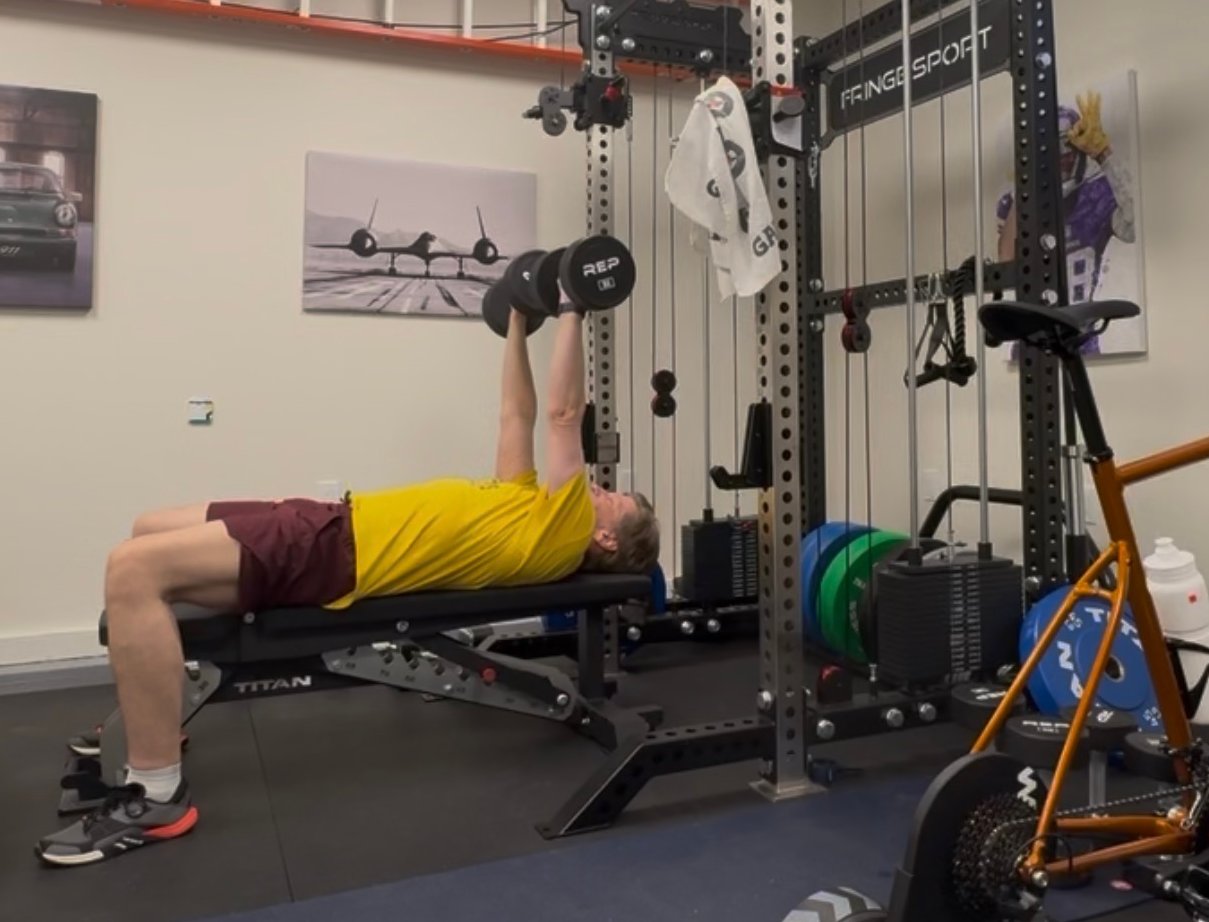 Gunnar, 53, doing a dumbbell bench press on a Titan bench in his garage gym, with a squat rack, bumper plates, and an indoor bike in the background