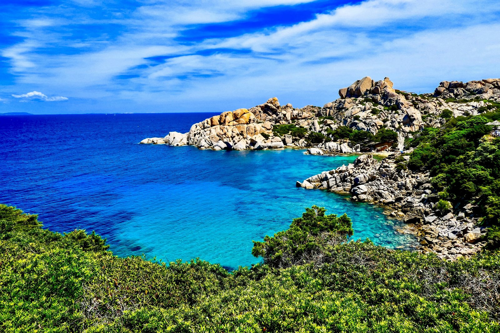 The granite coast at Capo Testa in northern Sardinia: turquoise shallows against a deep blue sea, weathered pale boulders, green scrub in the foreground under a bright sky.