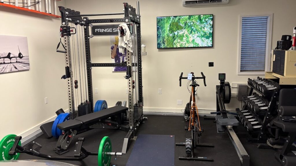 Home gym for men over 50: half rack with an adjustable bench and plates, a road bike on an indoor trainer, a rower, and a dumbbell rack.