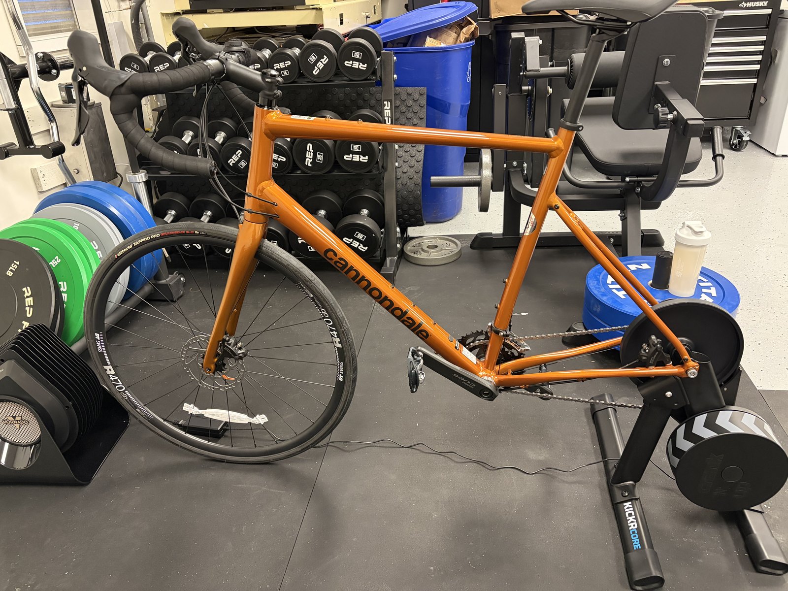 An orange Cannondale Synapse road bike mounted on a Wahoo KICKR Core trainer in a garage gym, with a rack of REP dumbbells behind it.